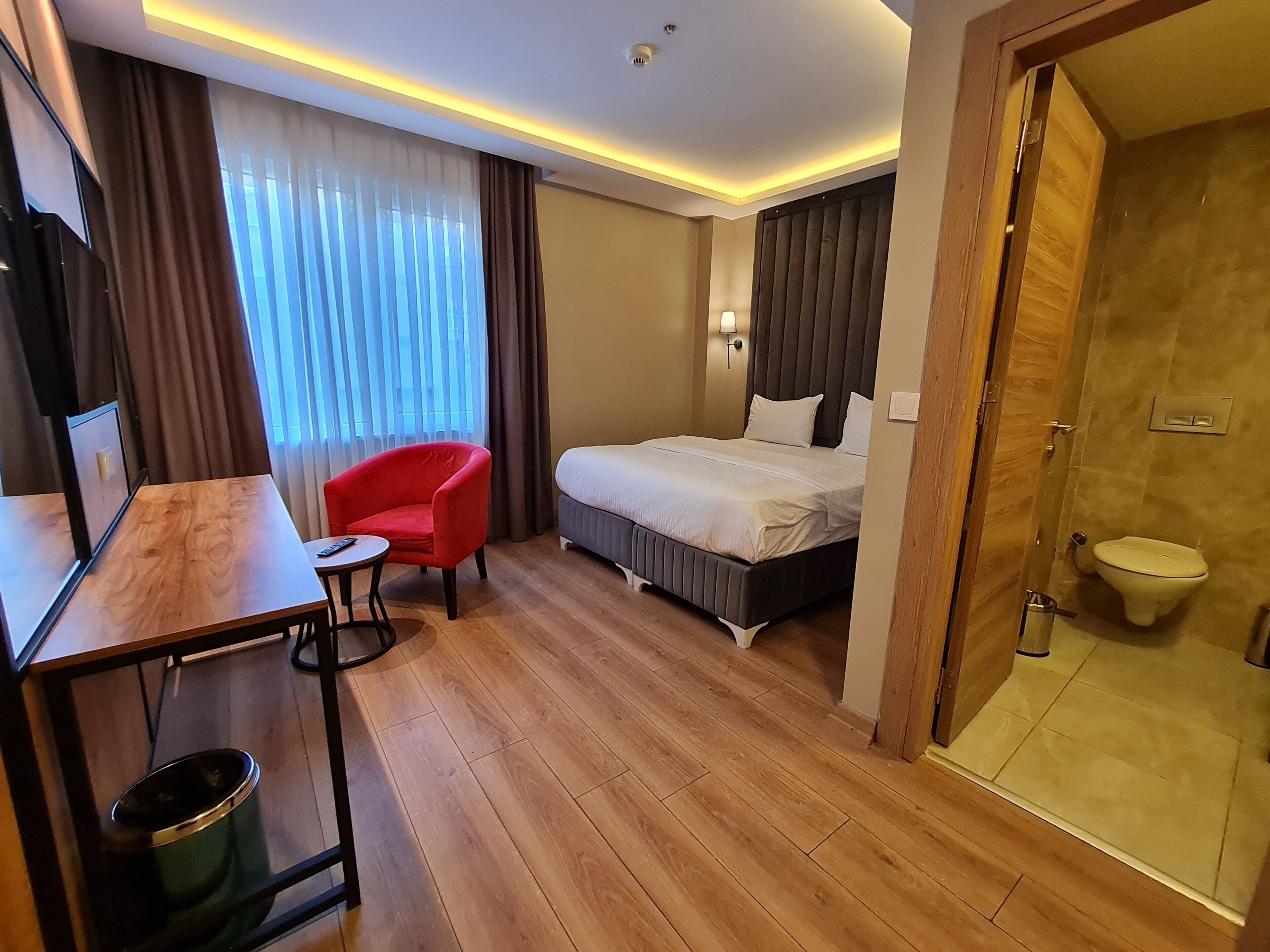 A hotel room with a wooden floor, a bed with a dark headboard, a red chair, a small table, a TV, and a bathroom with a toilet and shower.