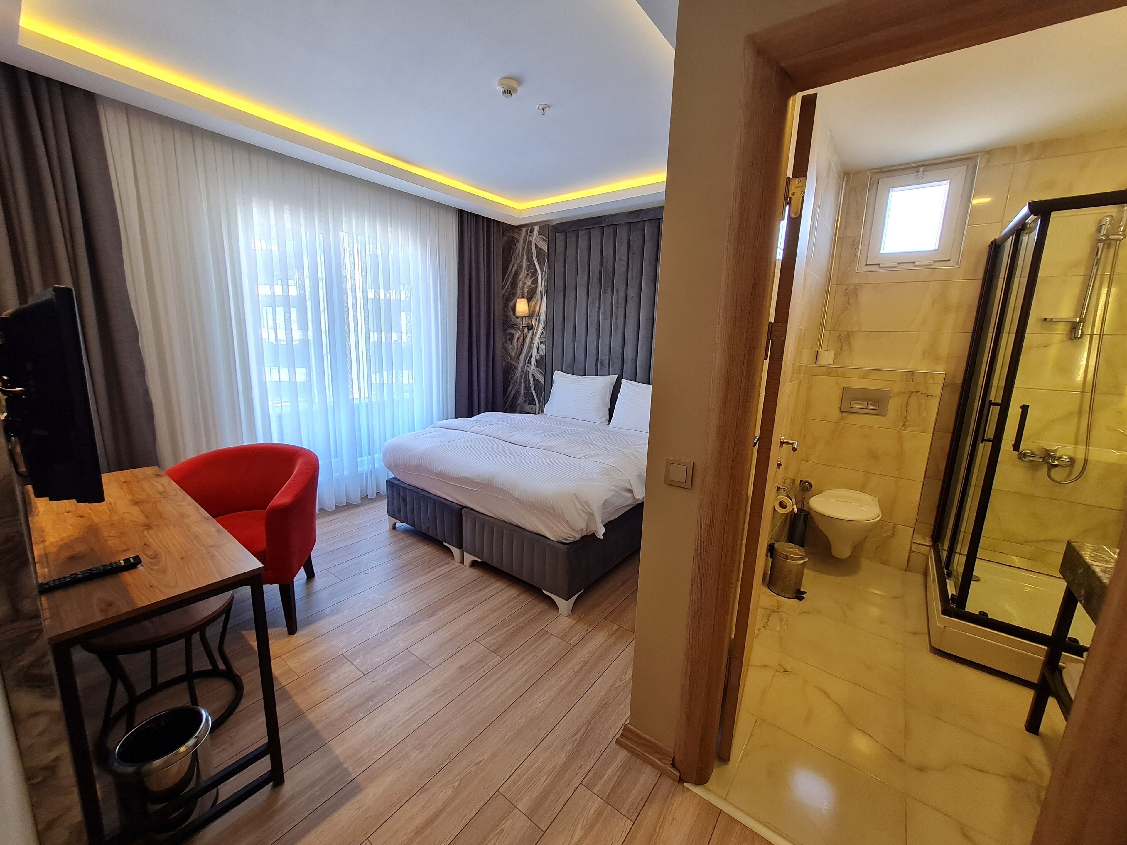 A hotel room with a bed, a desk, a red chair, a shower area, and a window with curtains.