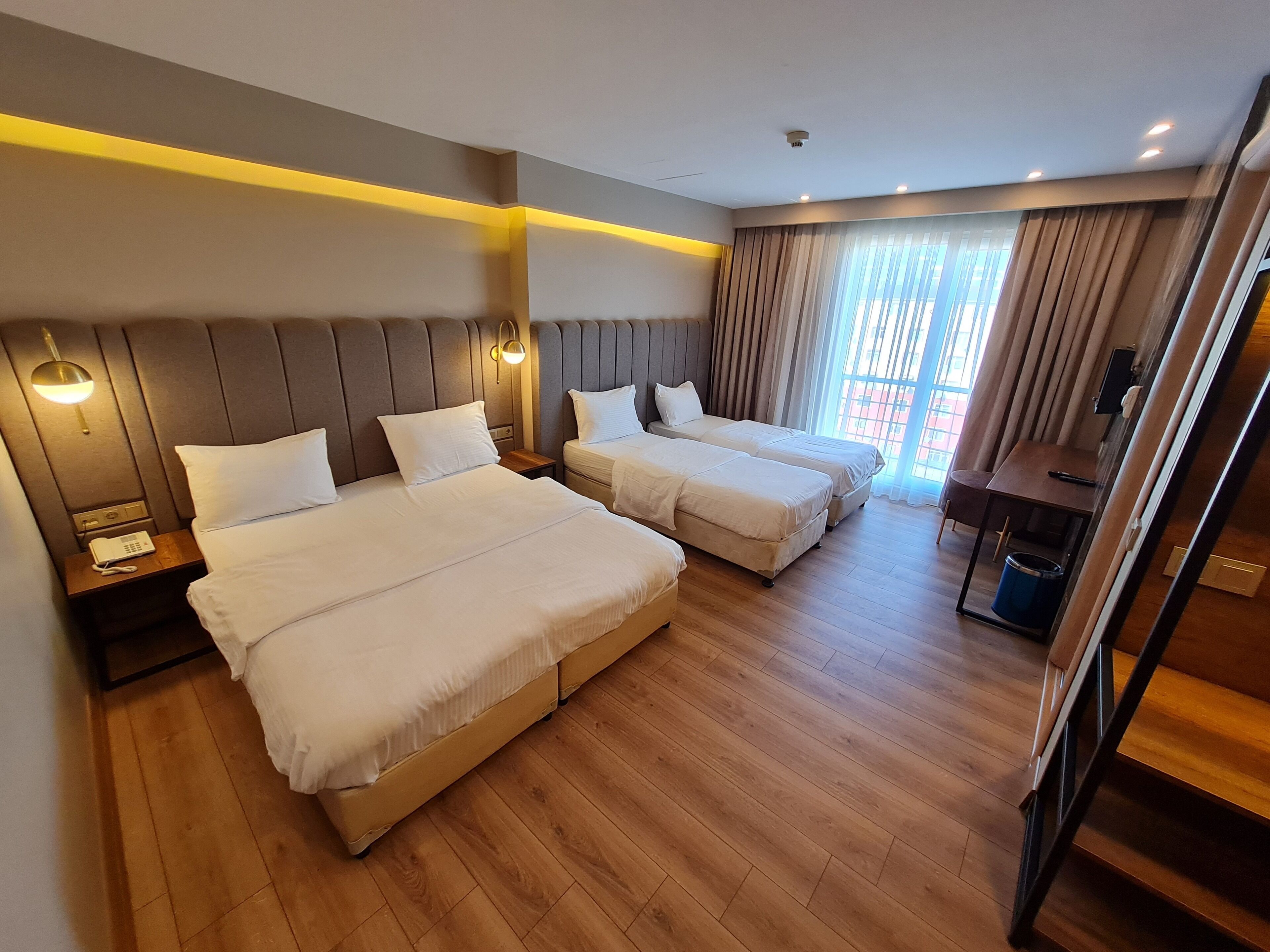 A hotel room with two beds, wooden flooring, a desk, and a window with curtains.