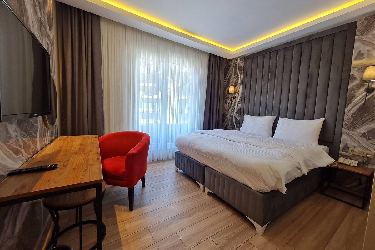 A modern hotel room with a large bed, a desk with a chair, a television, and a window with curtains.