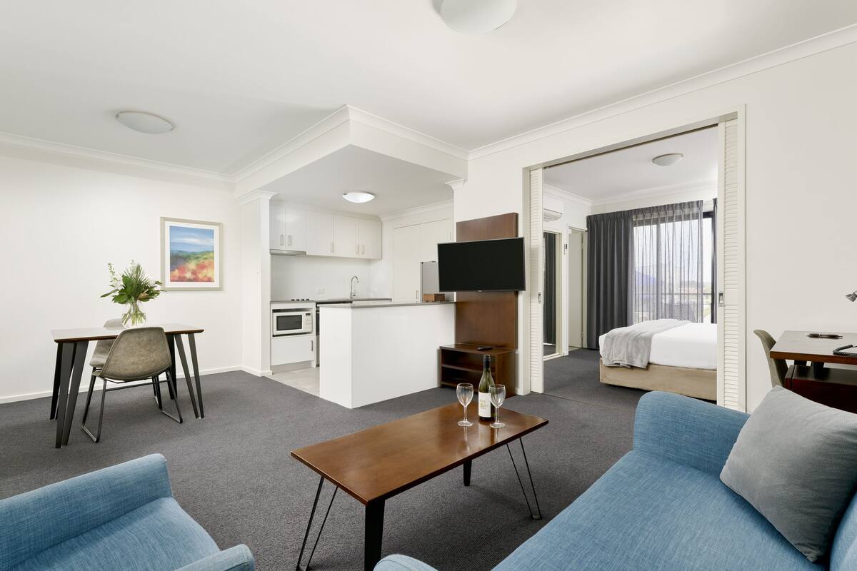 A hotel room with a kitchenette, a dining area, a living room with a sofa, and a bedroom with a bed and curtains.