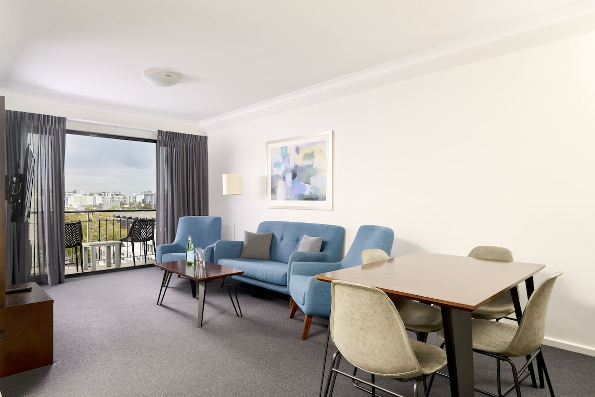 A hotel room with a blue sofa, a dining table with chairs, a small coffee table, a lamp, and a view of the city through large windows.