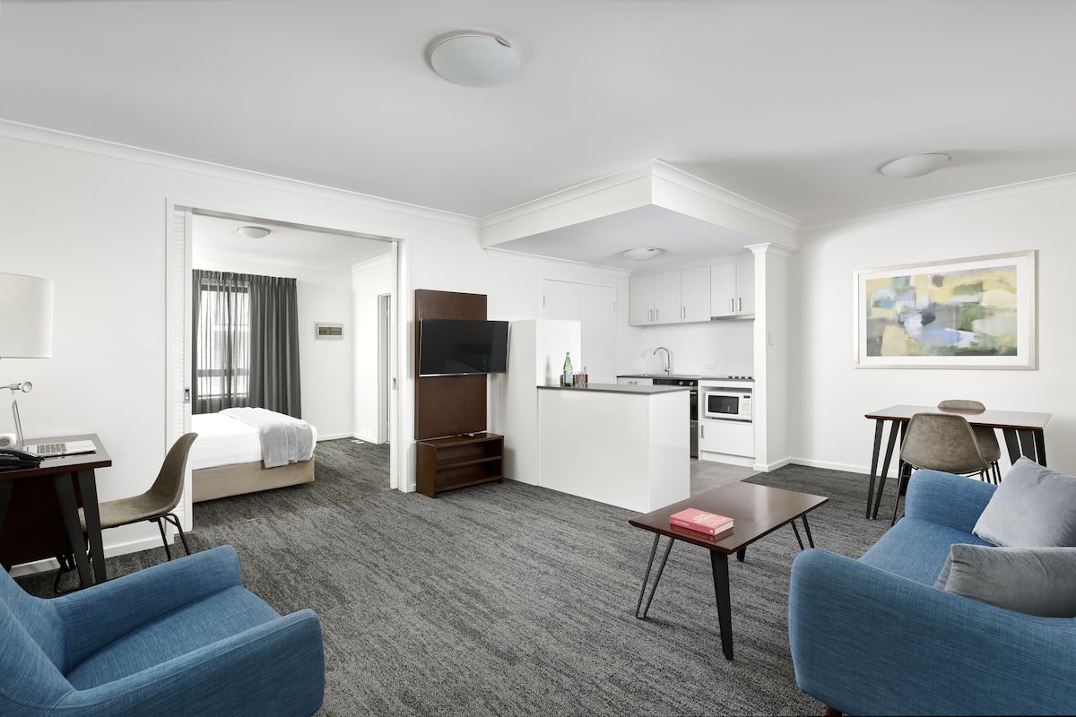 A hotel room with a bed, a desk, a chair, a sofa, a coffee table, a TV, a kitchenette, and a painting on the wall.