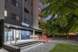 A modern building with a sign reading "QUEST" at the entrance, featuring a staircase, a glass door, and a well-lit pathway.
