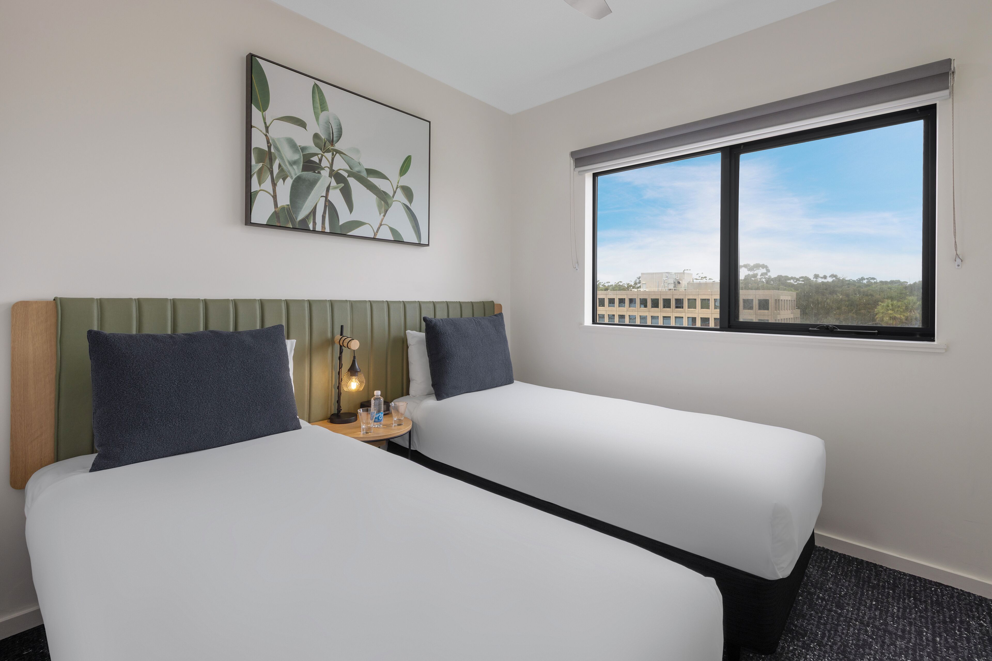 A hotel room with two beds, a small table with a lamp, a painting on the wall, and a window with a city view.