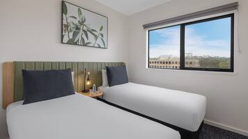 A hotel room with two beds, a small table with a lamp, a painting on the wall, and a window with a city view.