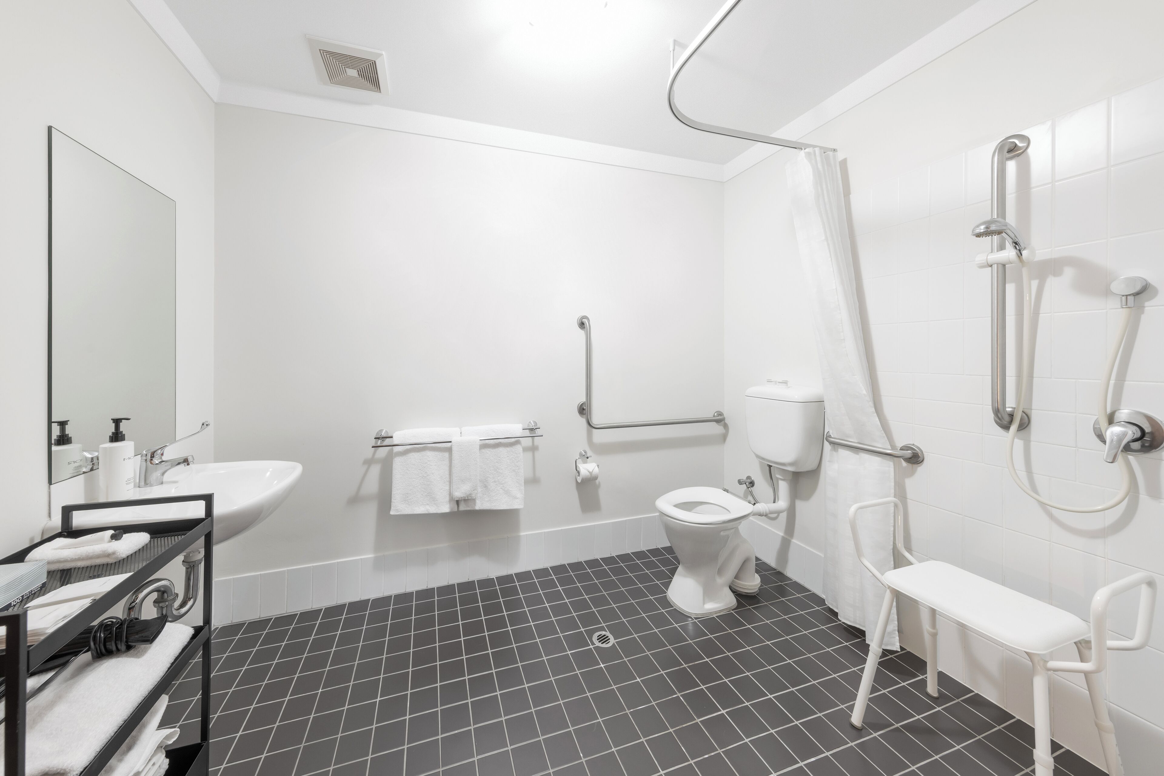 A modern bathroom with a white sink, toilet, and shower. It includes a bench and a handrail for accessibility.