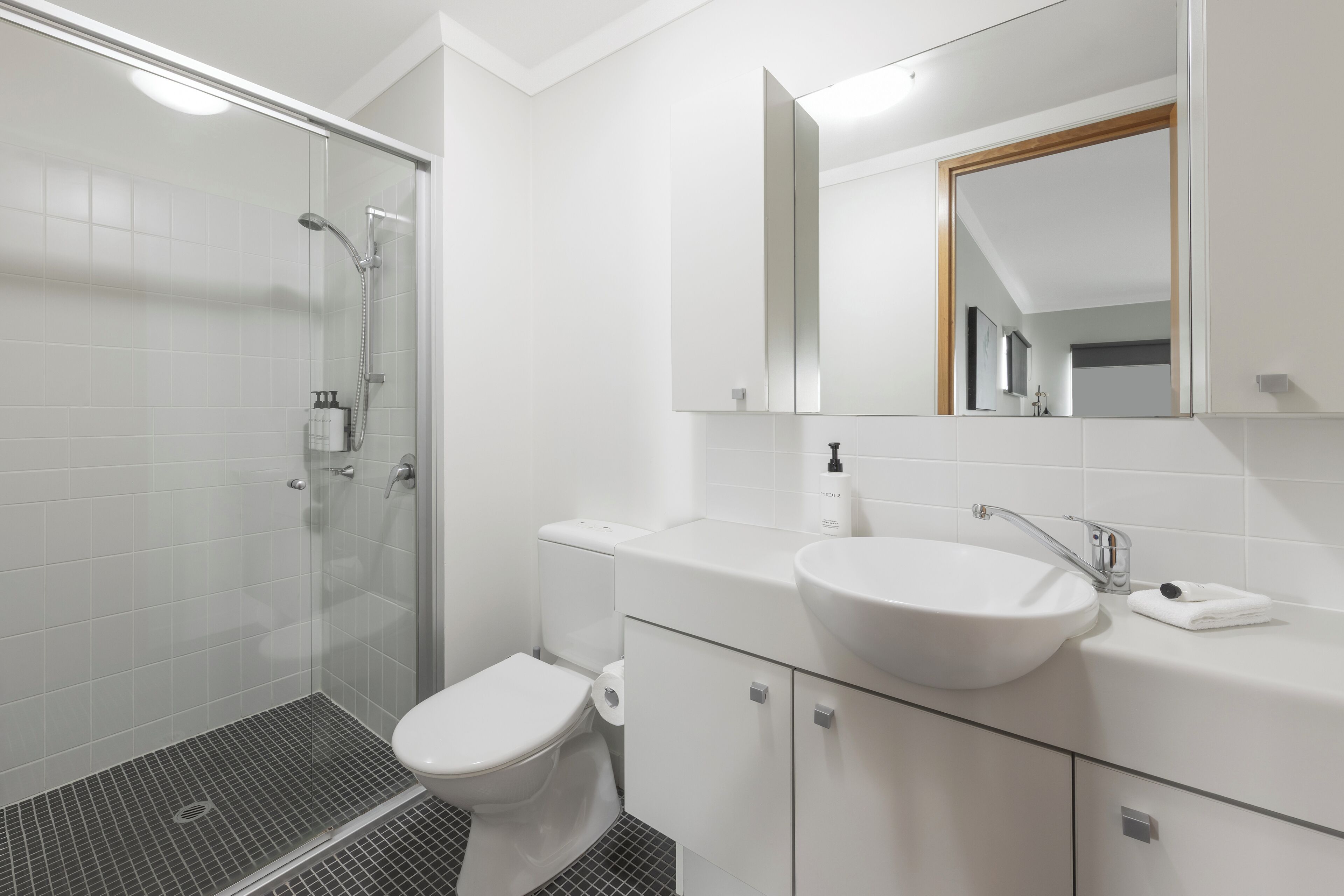 Un baño moderno con cabina de ducha de vidrio, inodoro, lavamanos con espejo y suelo de baldosas.