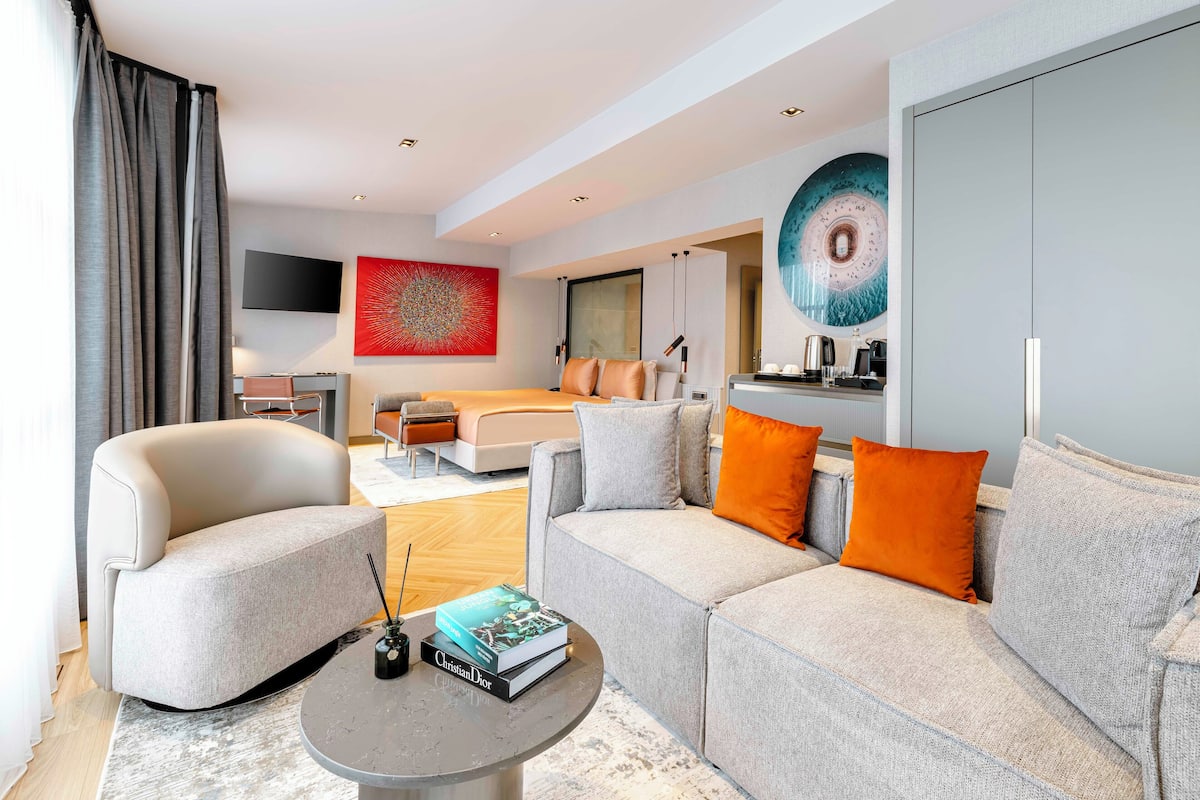 Quarto de hotel moderno com sofá cinzento, mesa de centro redonda com livros, televisão de tela plana, pintura abstrata grande e cama com almofadas laranja.