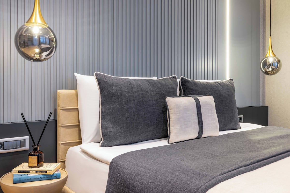 Quarto moderno com cama grande, roupa de cama cinzenta e duas luminárias pendentes.