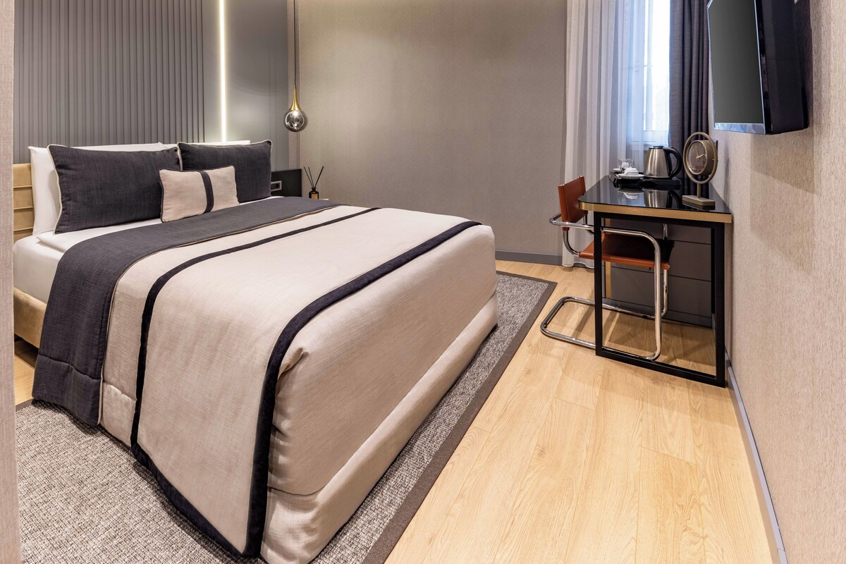 A modern hotel room with a large bed, a desk with a chair, a flat-screen TV, and a window with curtains.