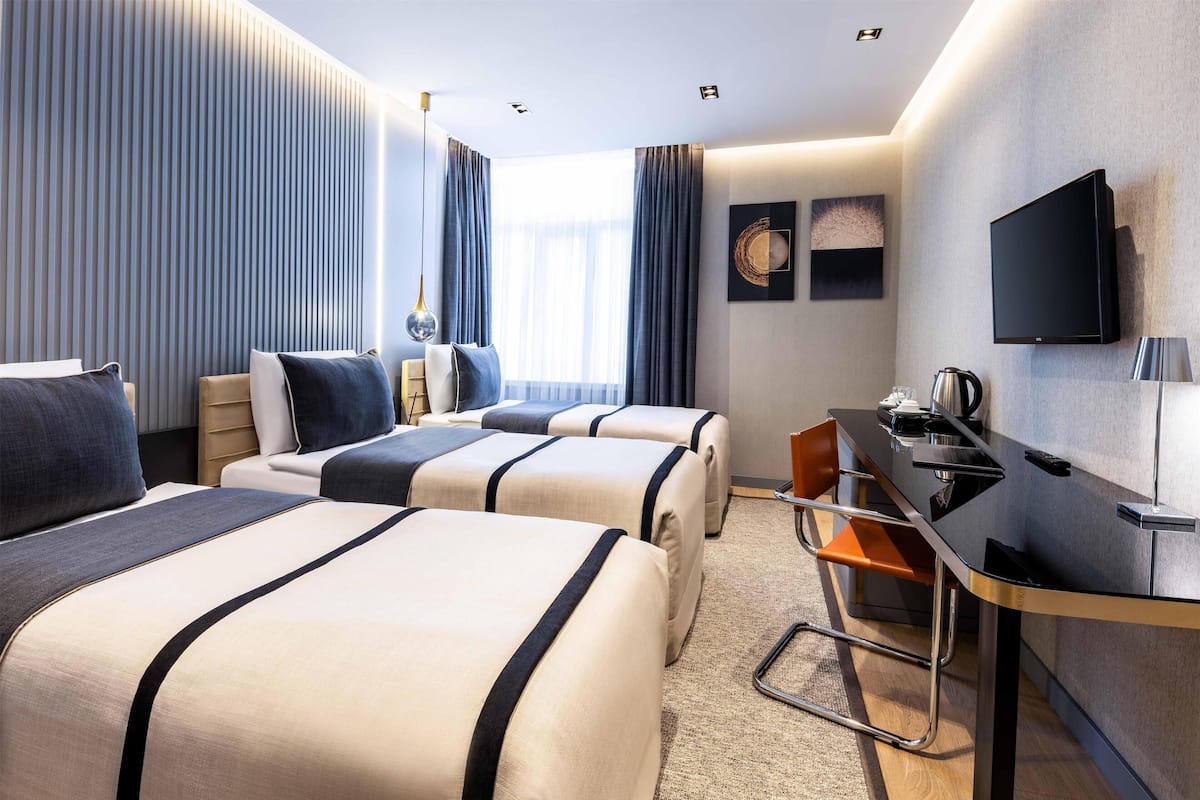 Quarto de hotel com duas camas, uma escrivaninha, uma televisão e uma janela com cortinas.