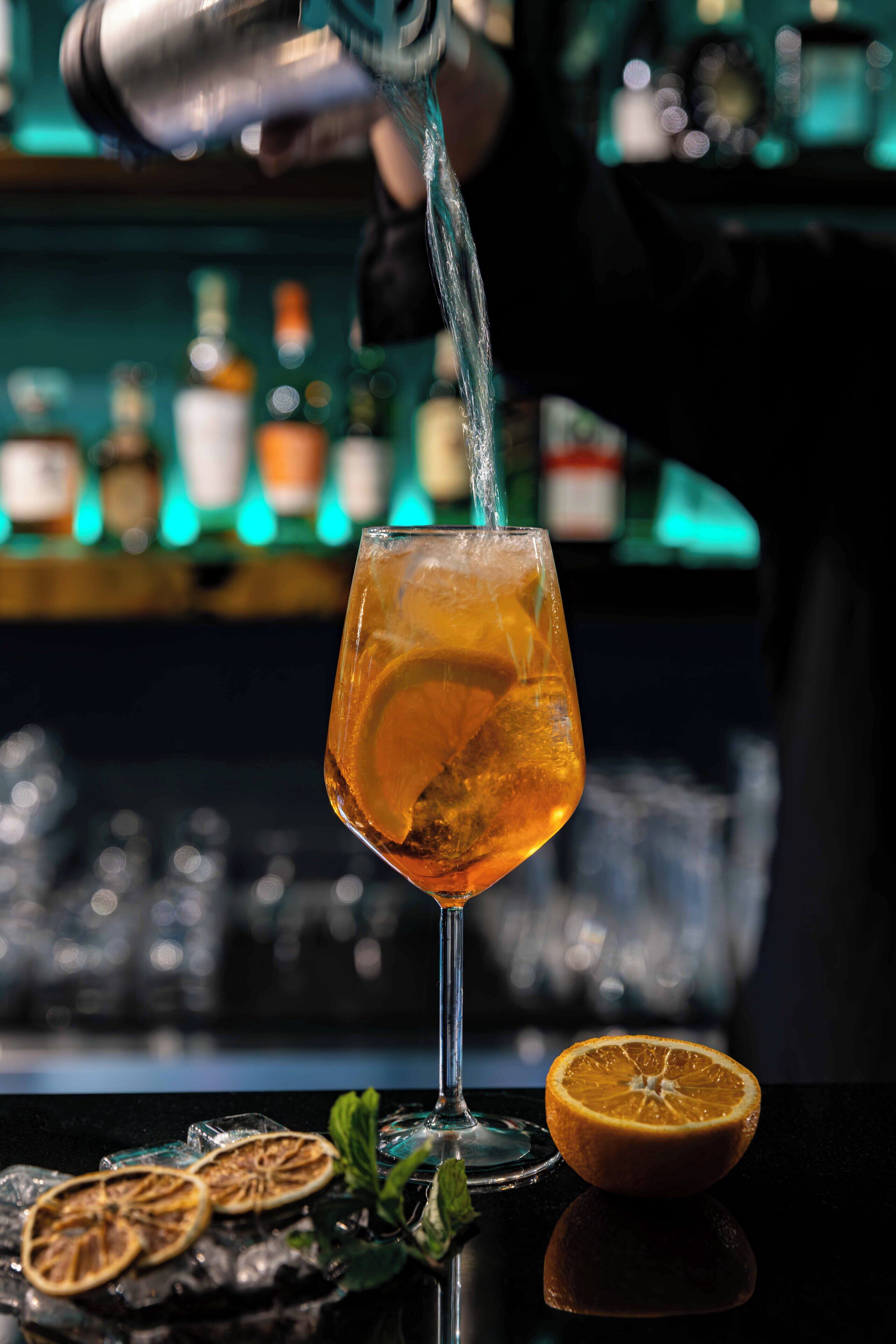 A cocktail being poured into a glass with an orange slice, ice, and mint leaves on a reflective surface.