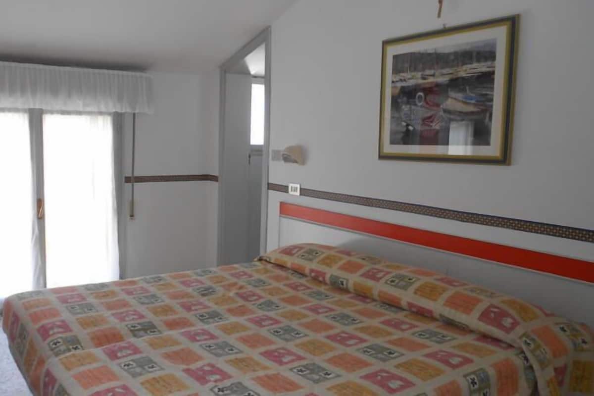 A bedroom with a bed, a window with curtains, a door, and a framed picture on the wall.