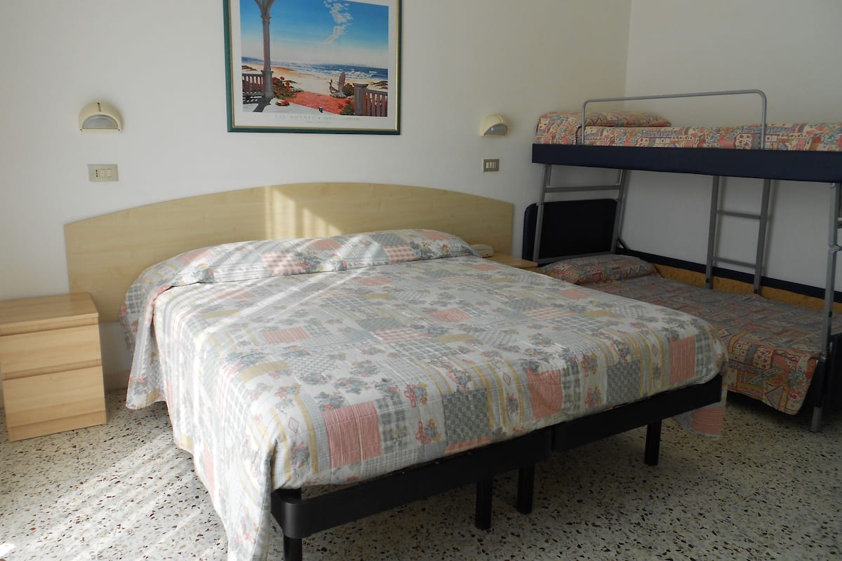 A bedroom with a single bed, a bunk bed, a wooden nightstand, a framed picture on the wall, and a tiled floor.