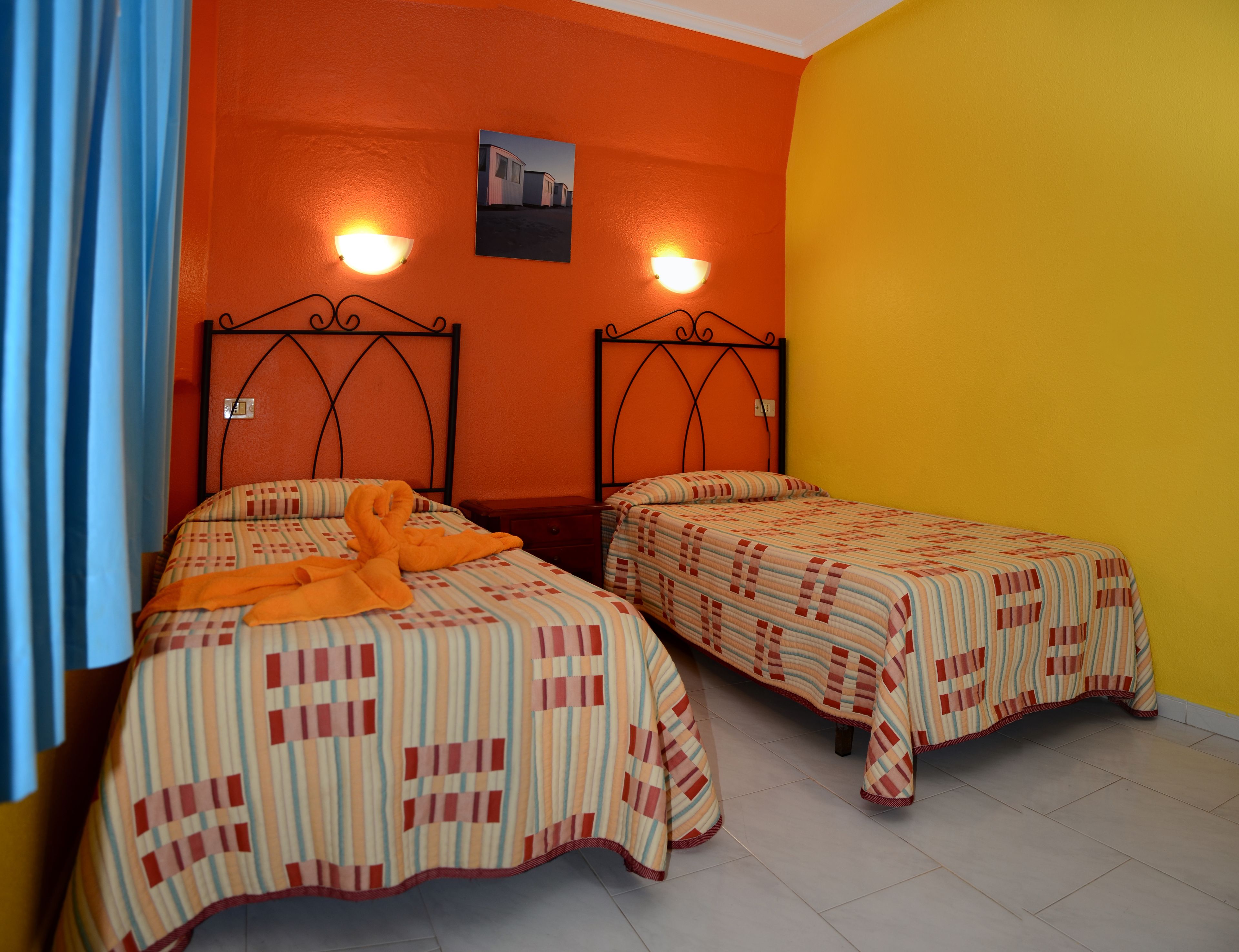 A room with two beds, each with striped bedding and a headboard. There are wall-mounted lights above the beds and a picture on the wall.