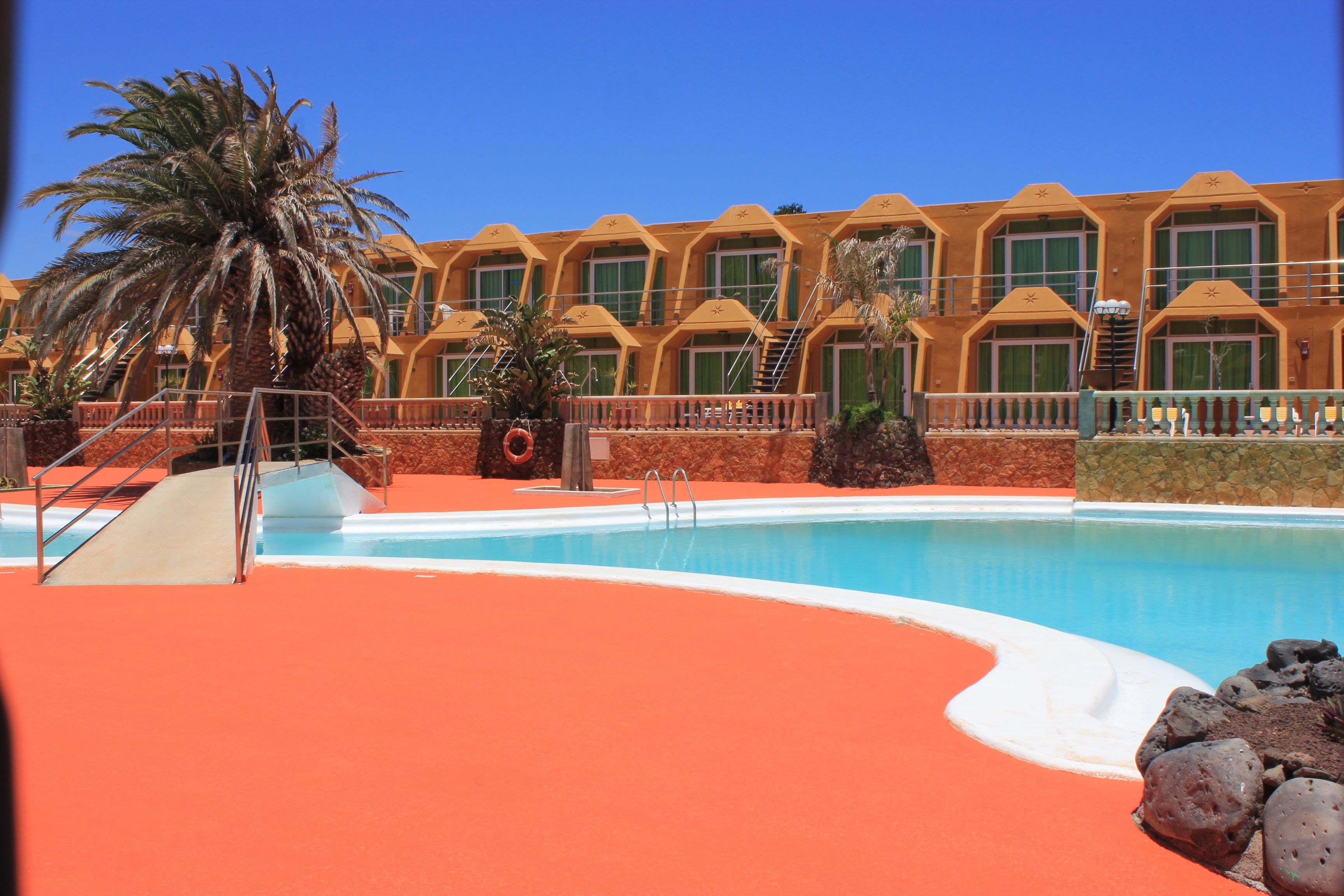 Un hotel con piscina, palmeras y una terraza de tejas rojas.