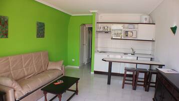 Una sala de estar con un sofá color beige, una mesa de centro y una cocina con encimera blanca, un fregadero y un microondas.
