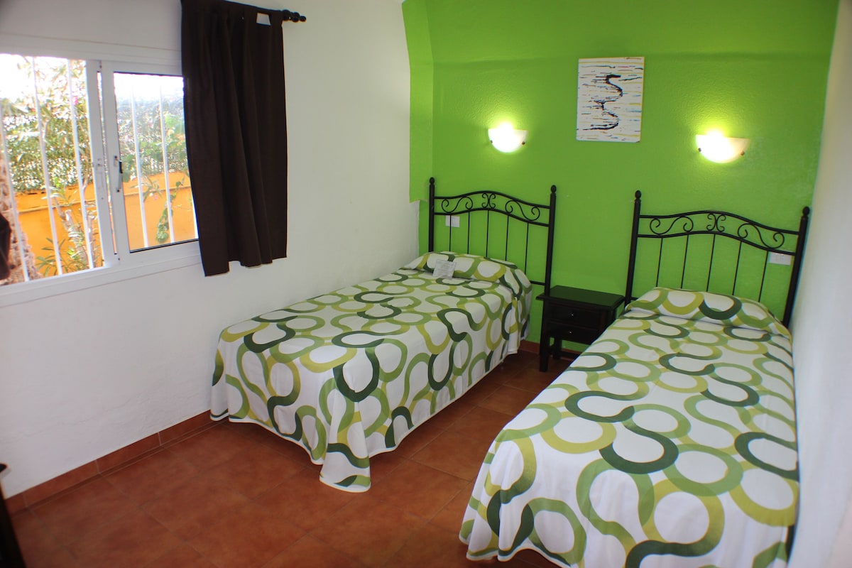 A room with two single beds, a window with curtains, and a green wall with two wall-mounted lights.