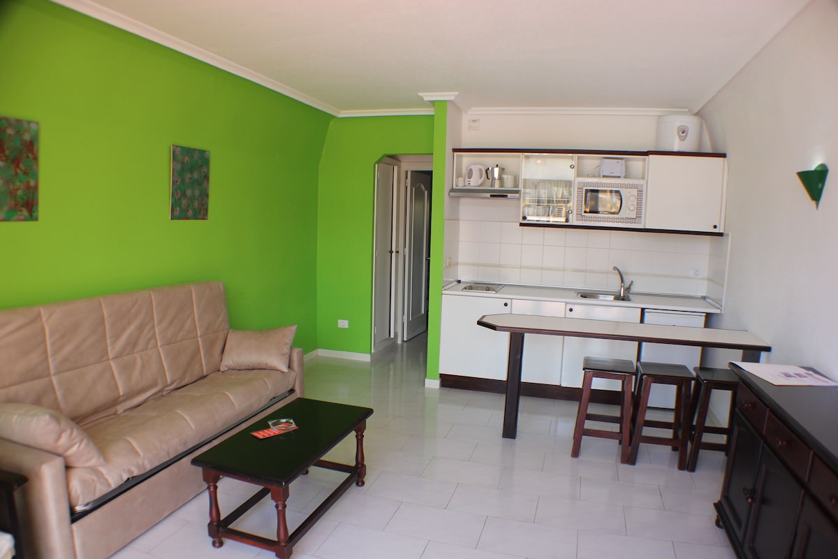 A living room with a beige sofa, a coffee table, and a kitchen area with a white countertop, a sink, and a microwave.