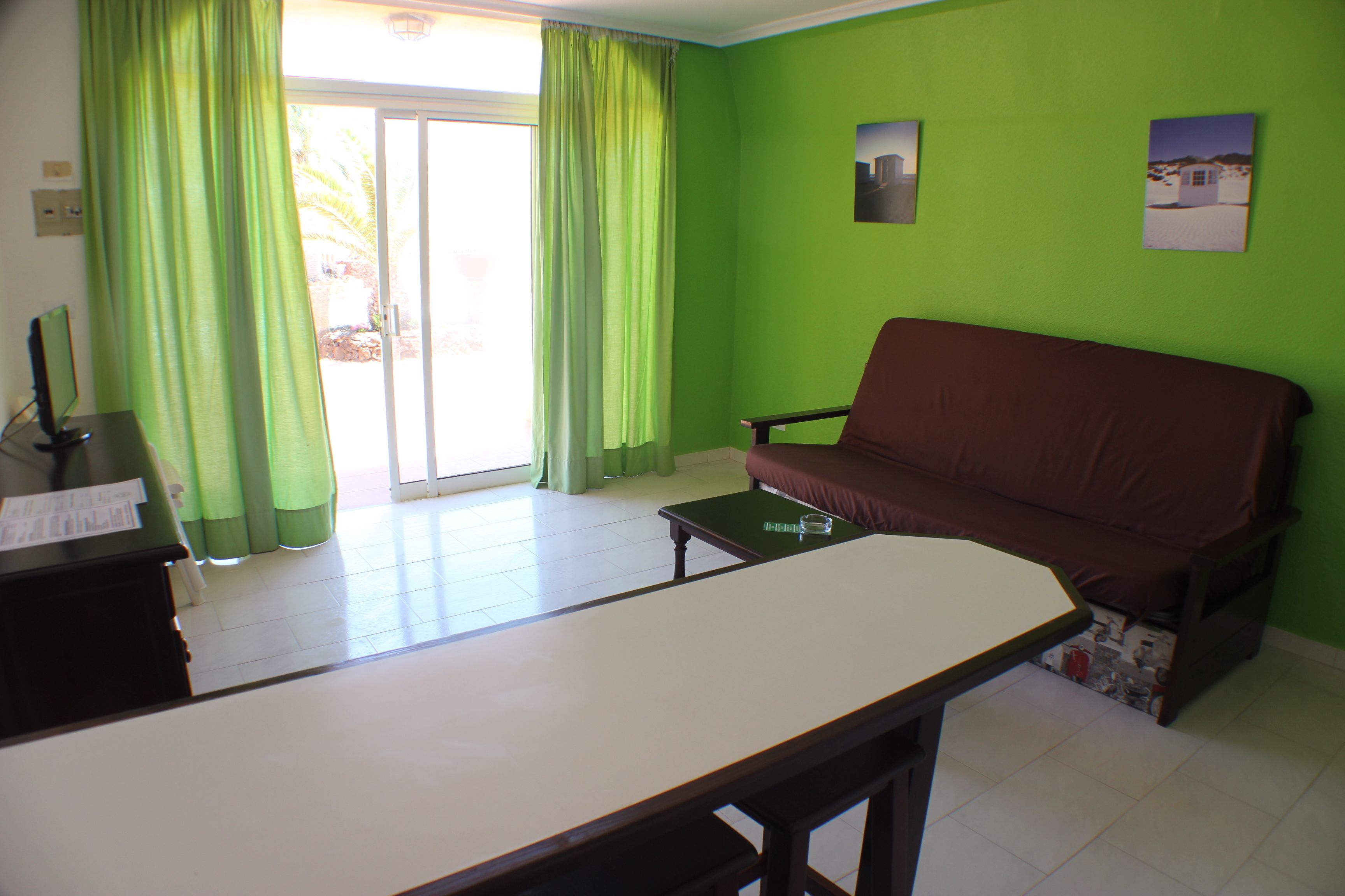 A room with a green wall, a brown sofa, a white table, and a sliding glass door with green curtains.