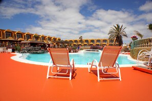 Un área de piscina con dos sillas de descanso, una piscina y una hilera de edificios amarillos al fondo.