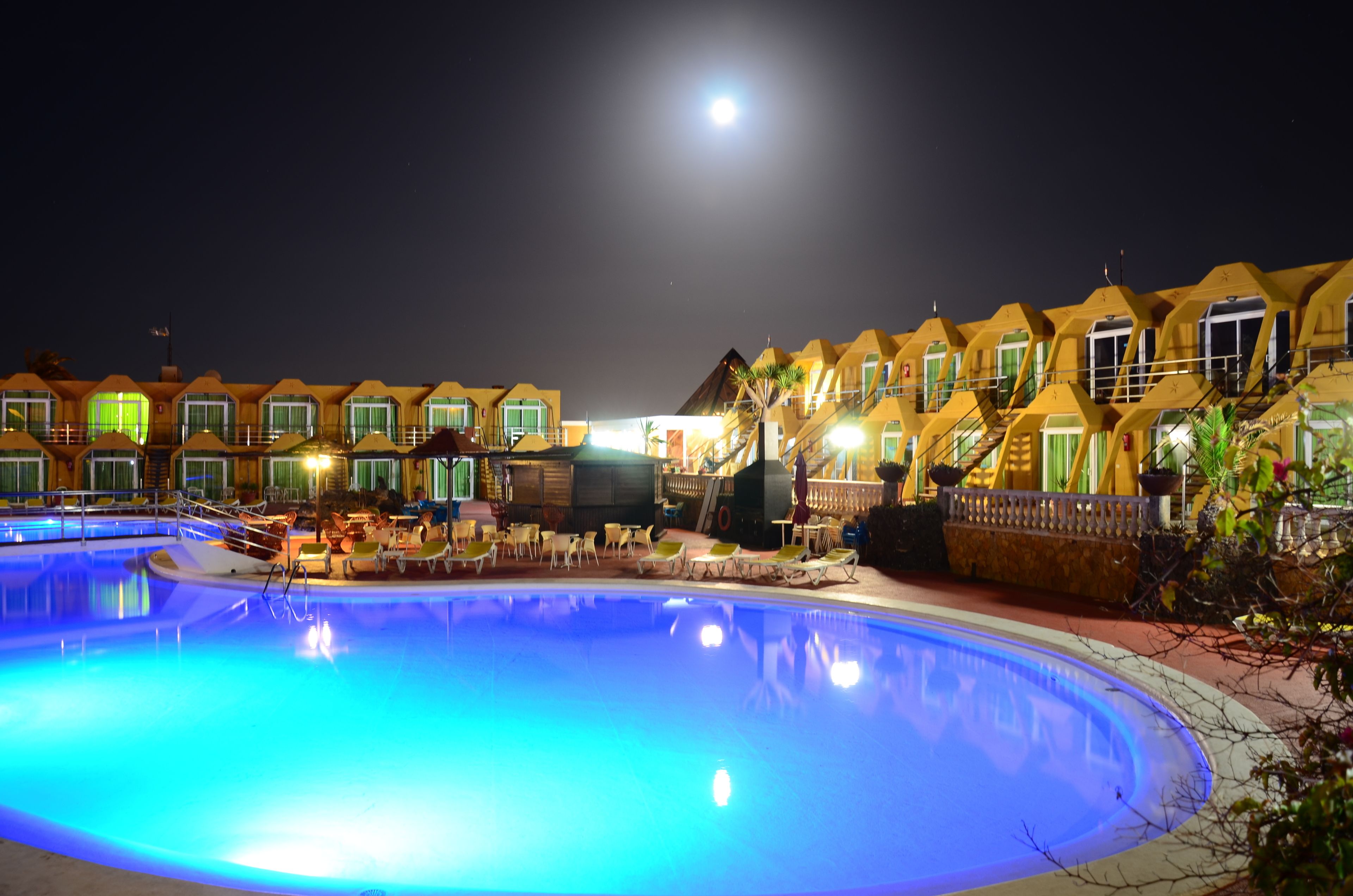 Zona de piscina con una luna en el cielo, rodeada de edificios amarillos con ventanas y una escalera.