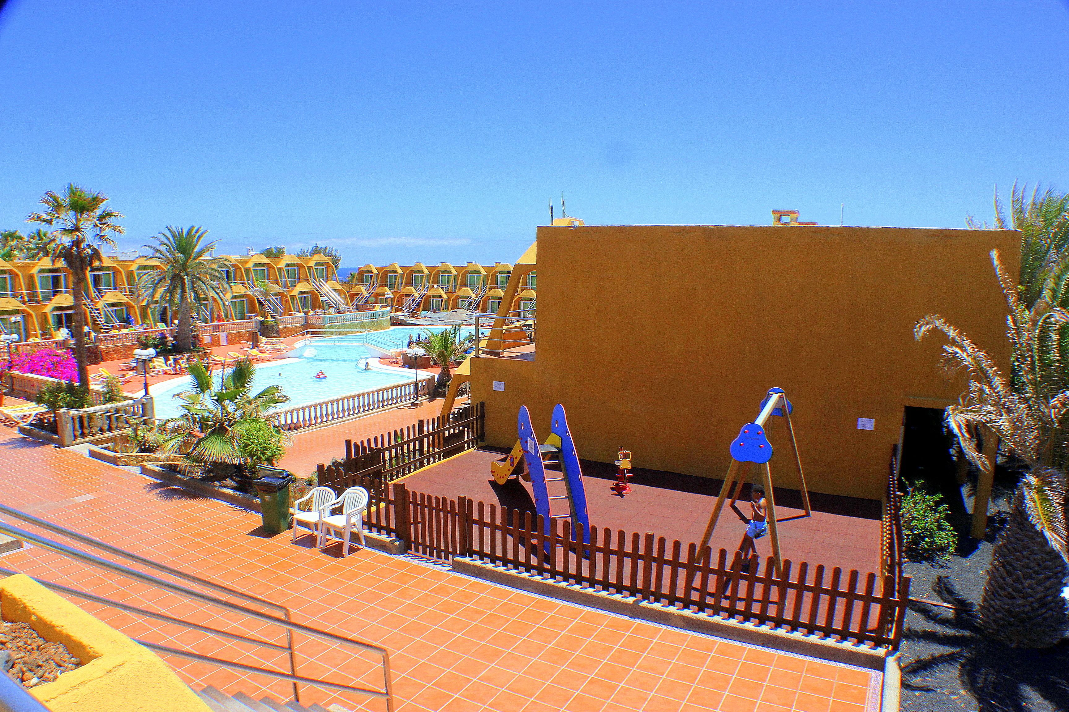 Ett semesterområde med en swimmingpool, palmer och en inh ägnad lekplats.