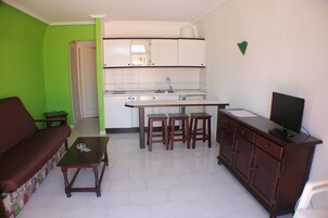 Un espacio habitable compacto con kitchenette, una zona de comedor con taburetes, un sofá y un televisor sobre un mueble.