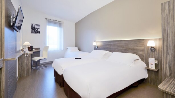 A hotel room with a large bed, a desk with a lamp, a TV mounted on the wall, and a window with curtains. - Kyriad Saint Chely d'Apcher - Aire de la Lozère (Albaret-Sainte-Marie)