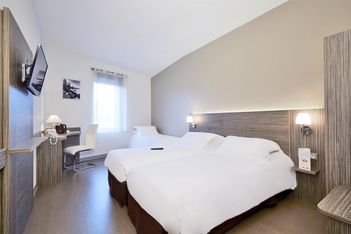 Habitación de hotel con una cama grande, un escritorio con lámpara, un televisor montado en la pared y una ventana con cortinas.