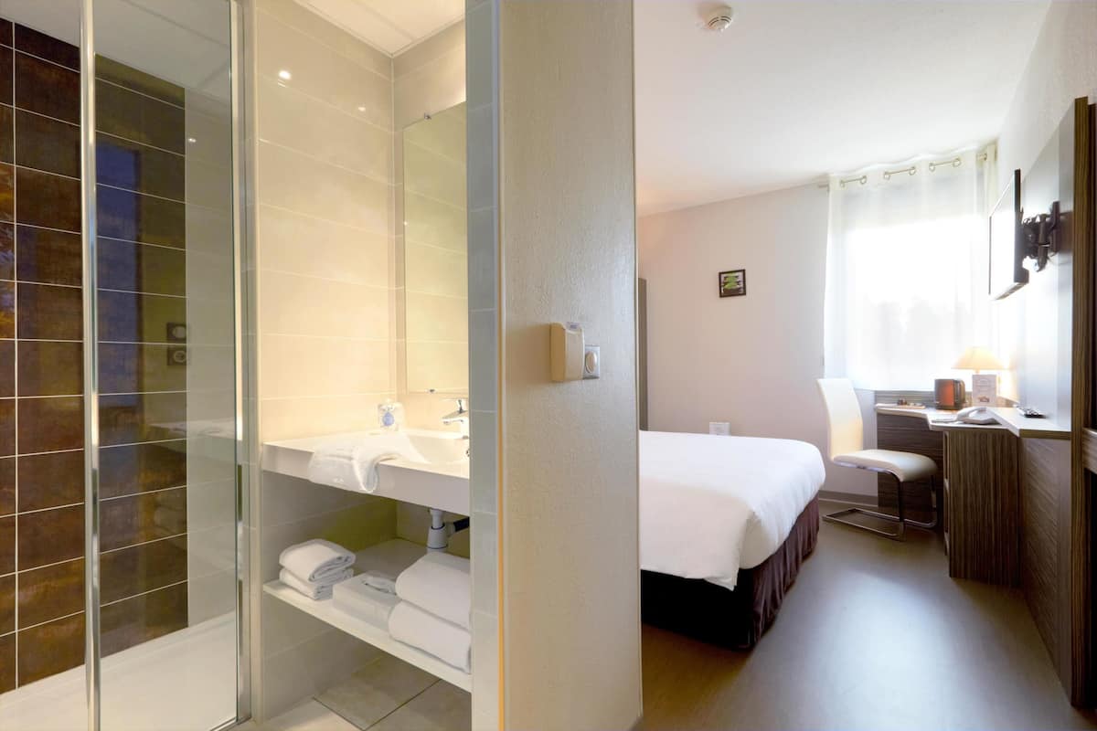 A hotel room with a glass shower enclosure, a bed, a desk with a chair, and a small table with a lamp.