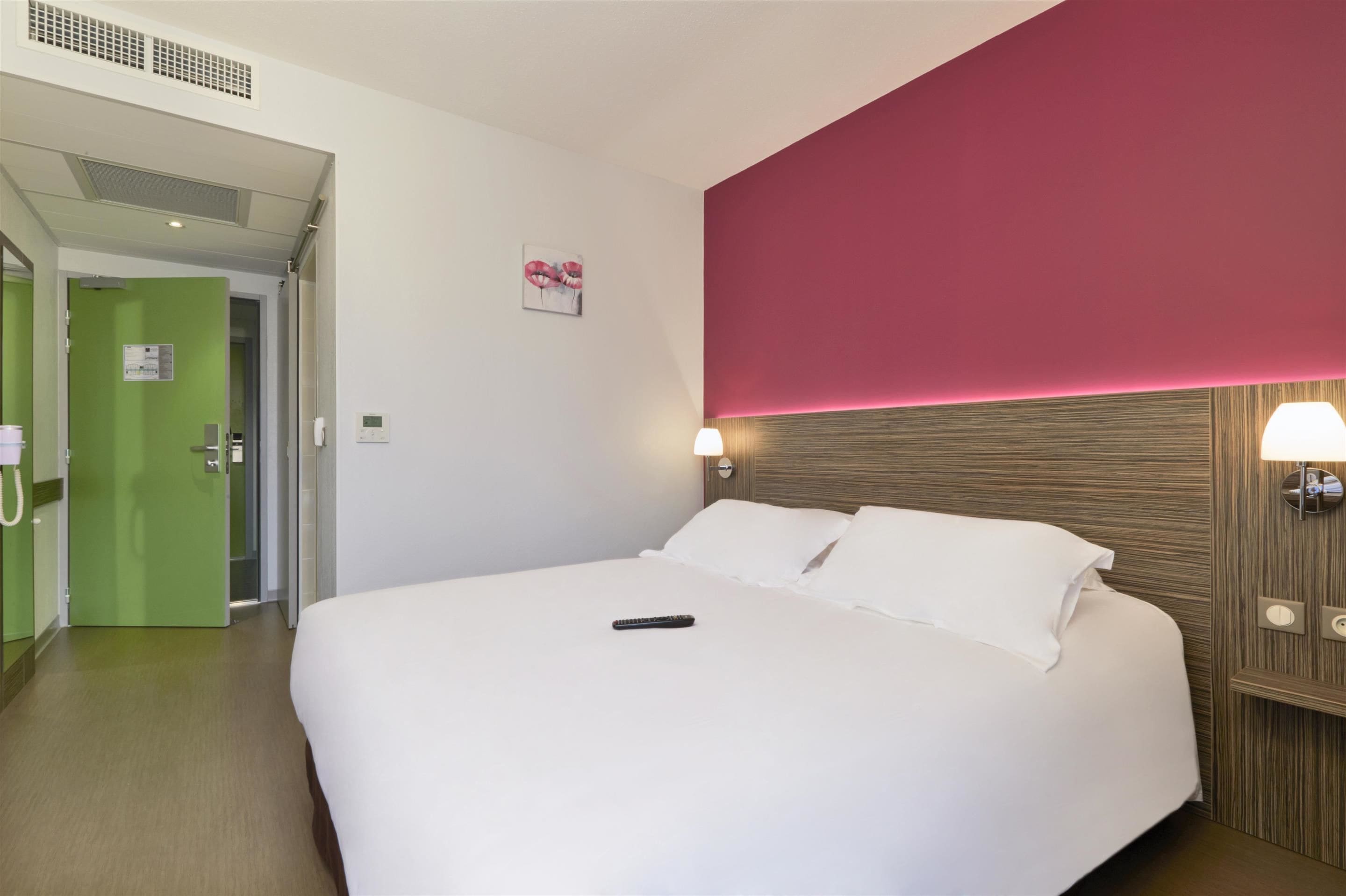 A hotel room with a large bed, a green door, a red accent wall, and a wall-mounted lamp.