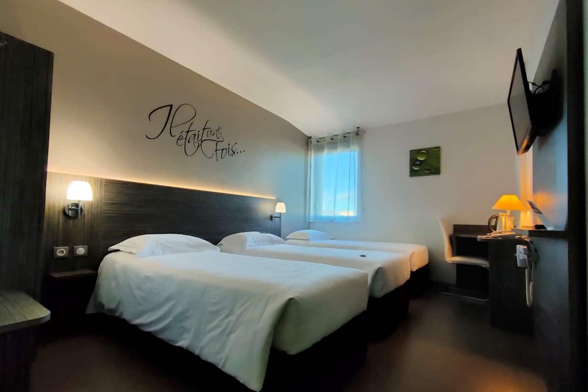 A hotel room with two beds, a desk with a lamp, a TV mounted on the wall, and a window with curtains.