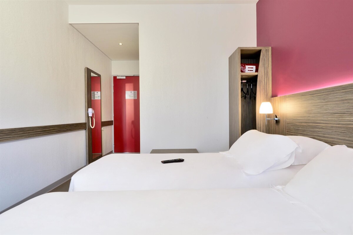 Una habitación de hotel con cama, mesita de noche con lámpara, una puerta roja y cabecero de madera.