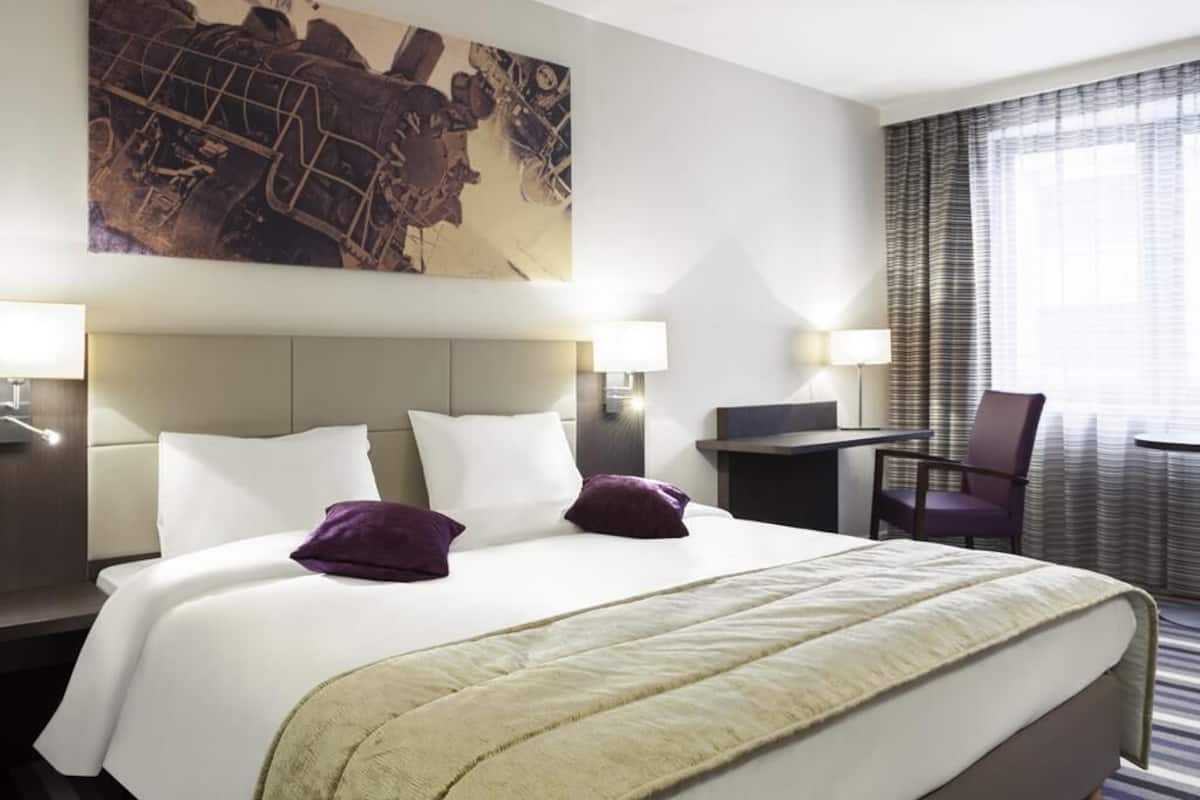 A hotel room with a large bed, two bedside tables, a desk, a chair, and a large window with curtains.