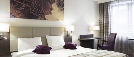 Une chambre d’hôtel avec un grand lit, deux tables de chevet, un bureau, une chaise et une grande fenêtre avec des rideaux.