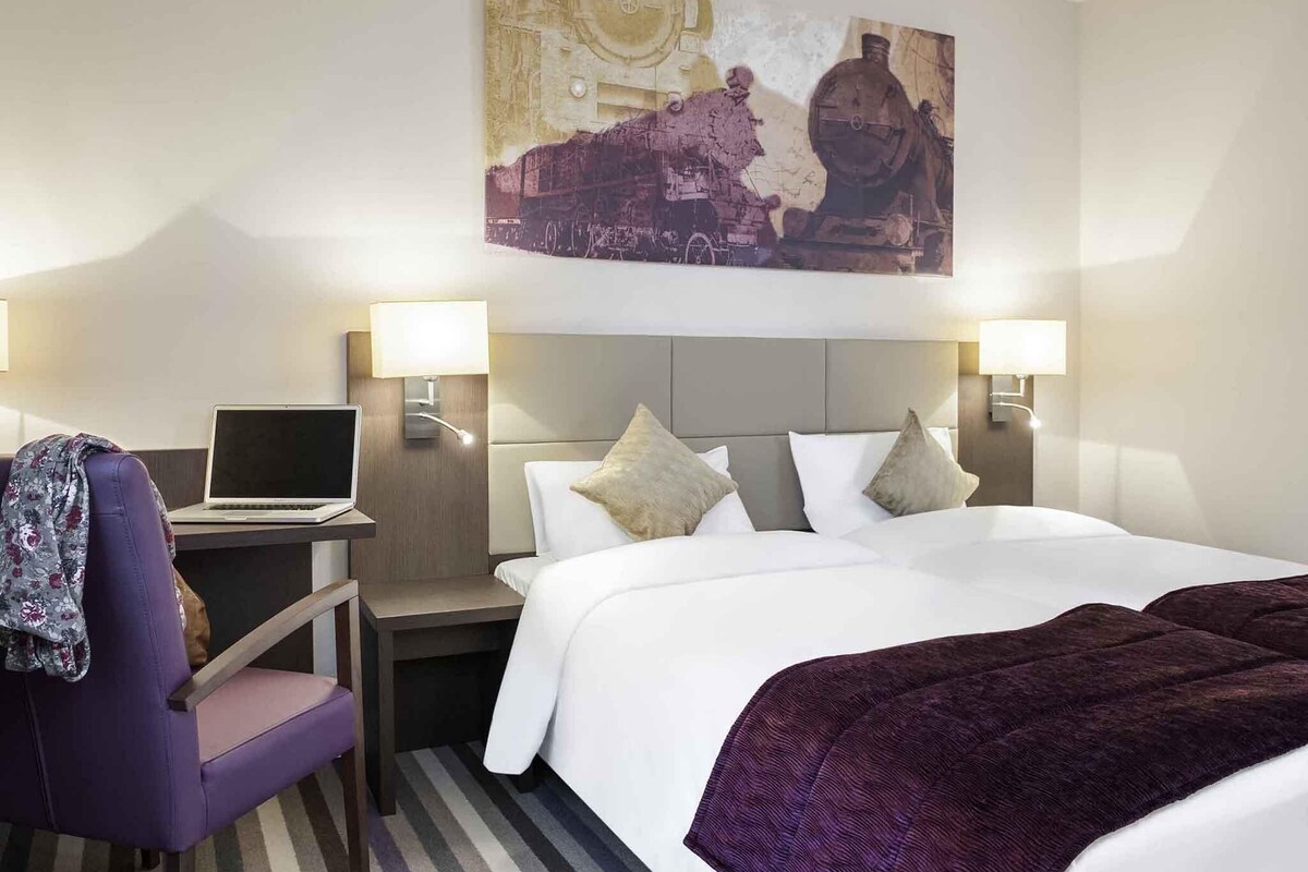 A hotel room with a bed, two bedside lamps, a laptop on a desk, a chair, and a large painting on the wall.