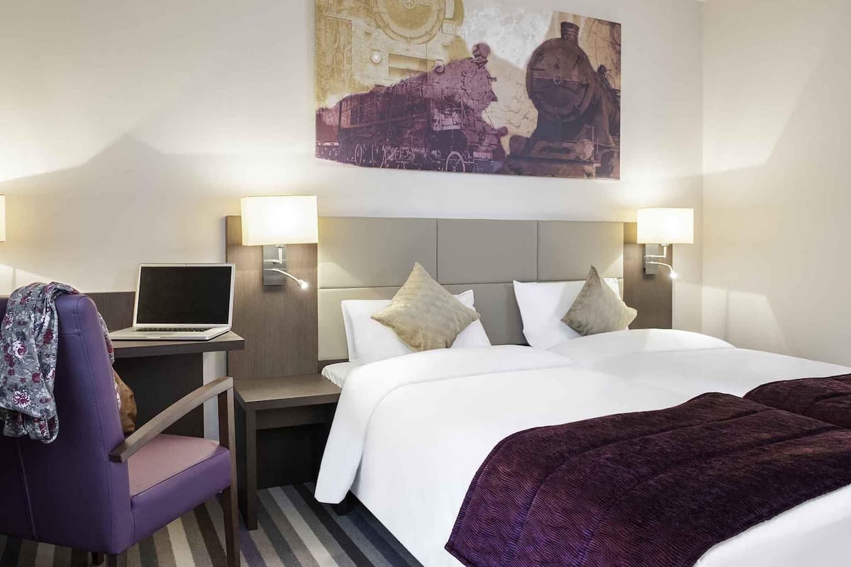 A hotel room with a bed, two bedside lamps, a laptop on a desk, a chair, and a large painting on the wall.