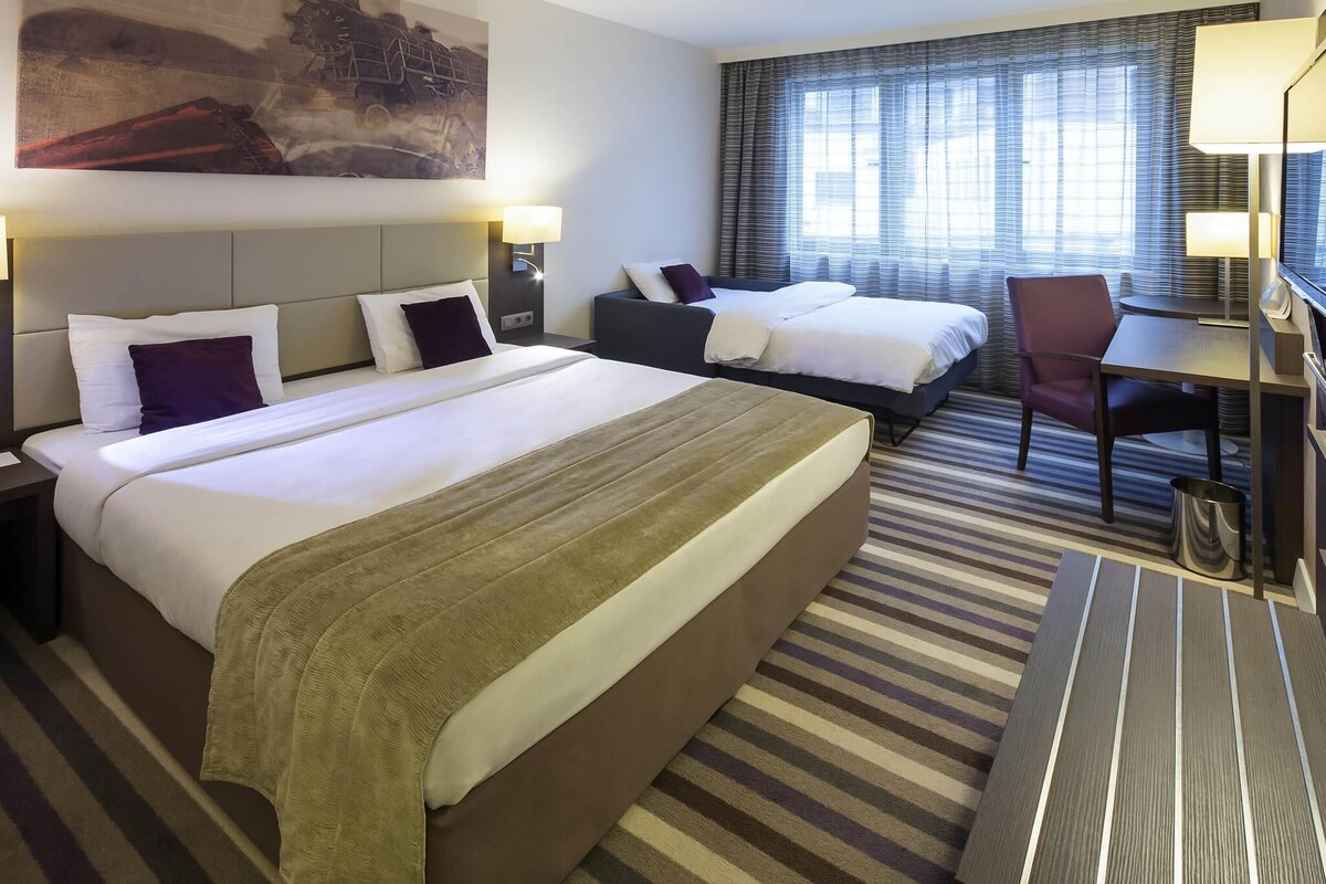 A hotel room with two beds, a desk, a chair, and a large painting on the wall.