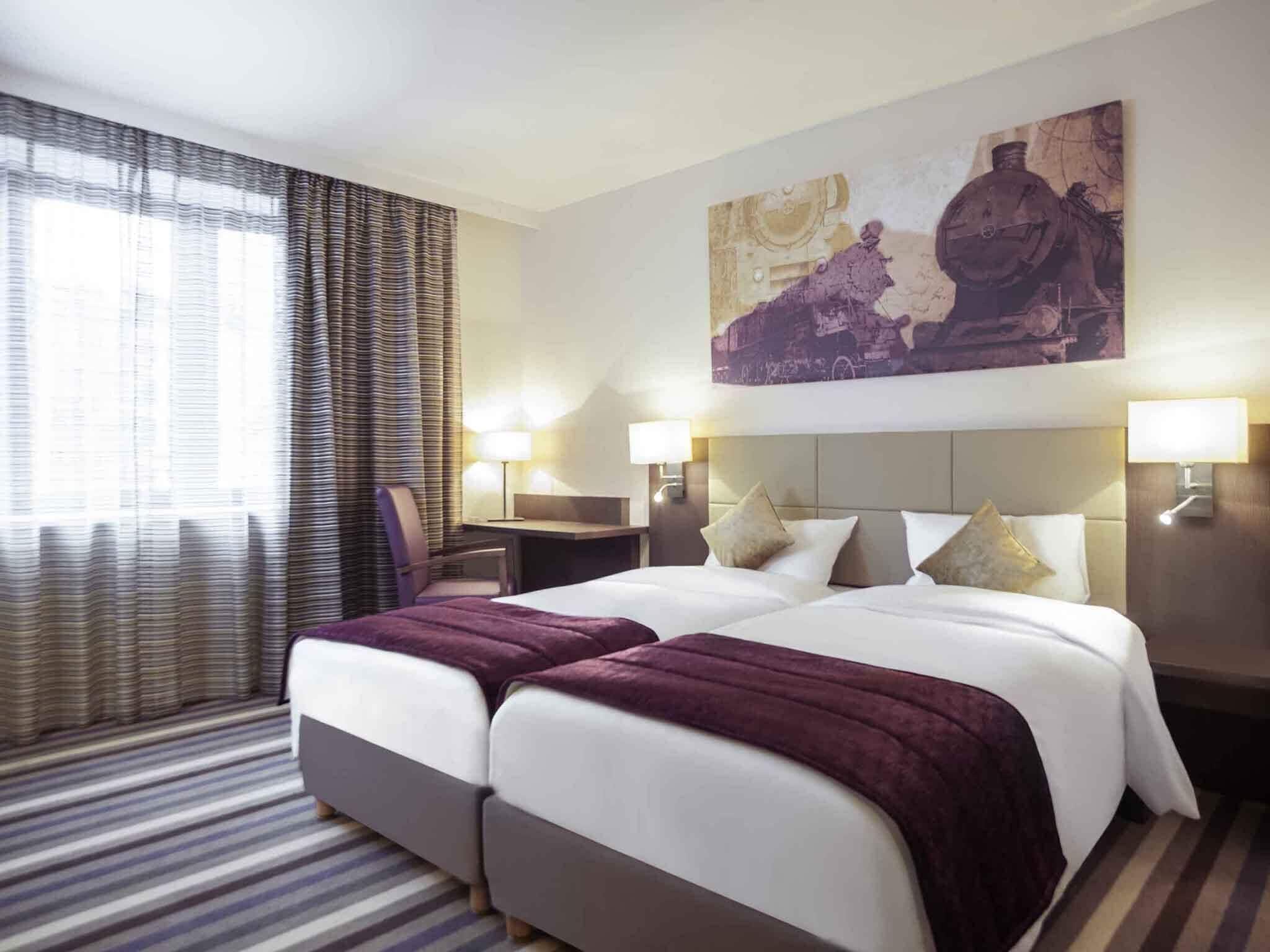 A hotel room with a double bed, two bedside tables, a desk, a chair, a large painting, and a window with curtains.