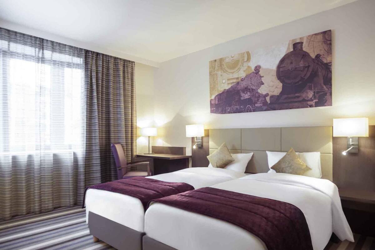 A hotel room with a double bed, two bedside tables, a desk, a chair, a large painting, and a window with curtains.