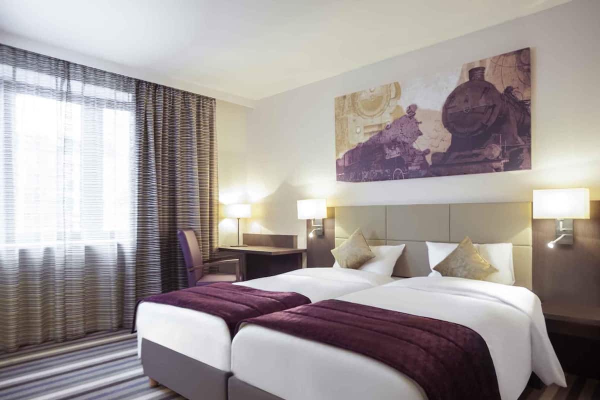 A hotel room with a double bed, two bedside tables, a desk, a chair, a large painting, and a window with curtains.