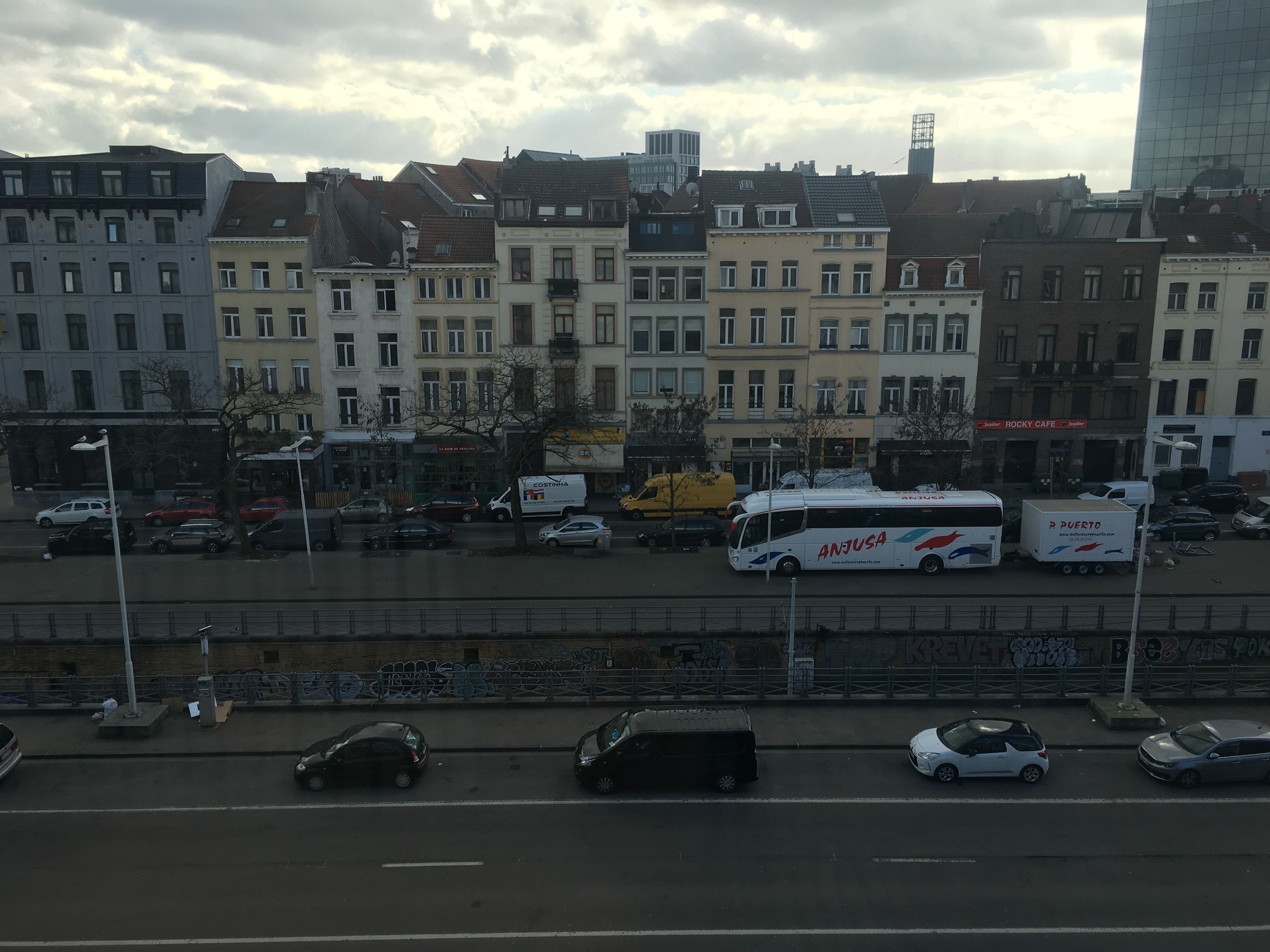 A street view with parked cars, a bus, and buildings with various storefronts.