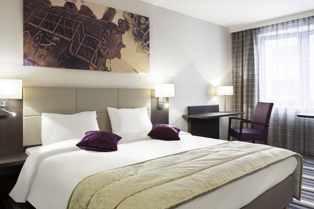 A hotel room with a large bed, two bedside tables with lamps, a desk, a chair, and a large painting on the wall.