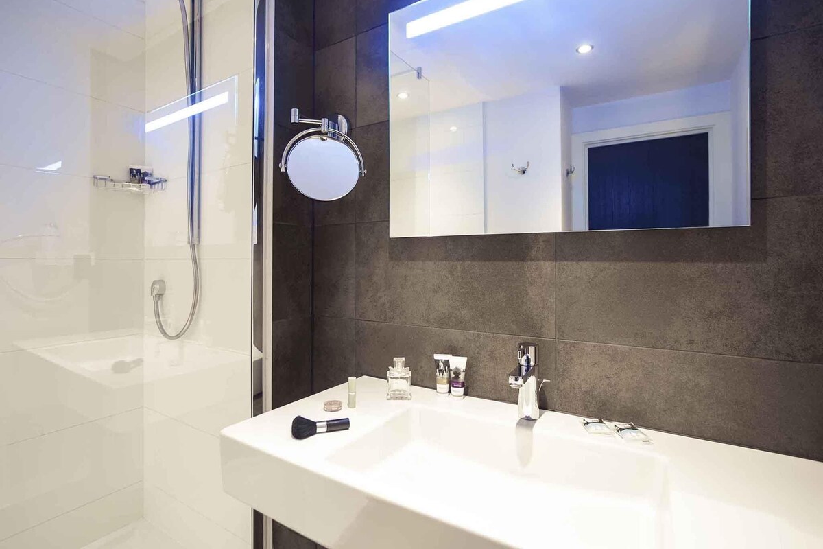 A modern bathroom with a glass shower enclosure, a white sink with two faucets, and a large mirror above the sink.