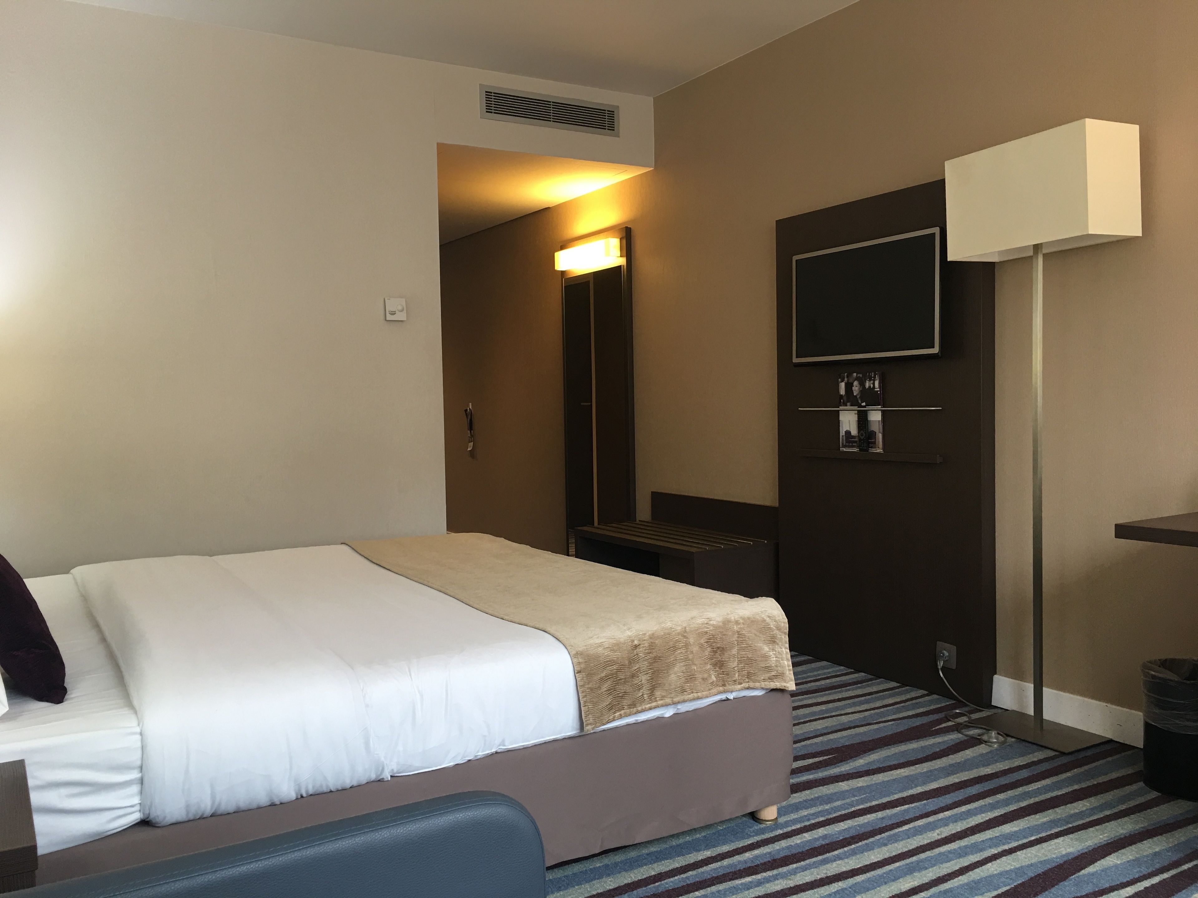 A hotel room with a bed, a television mounted on the wall, a desk, and a trash bin.