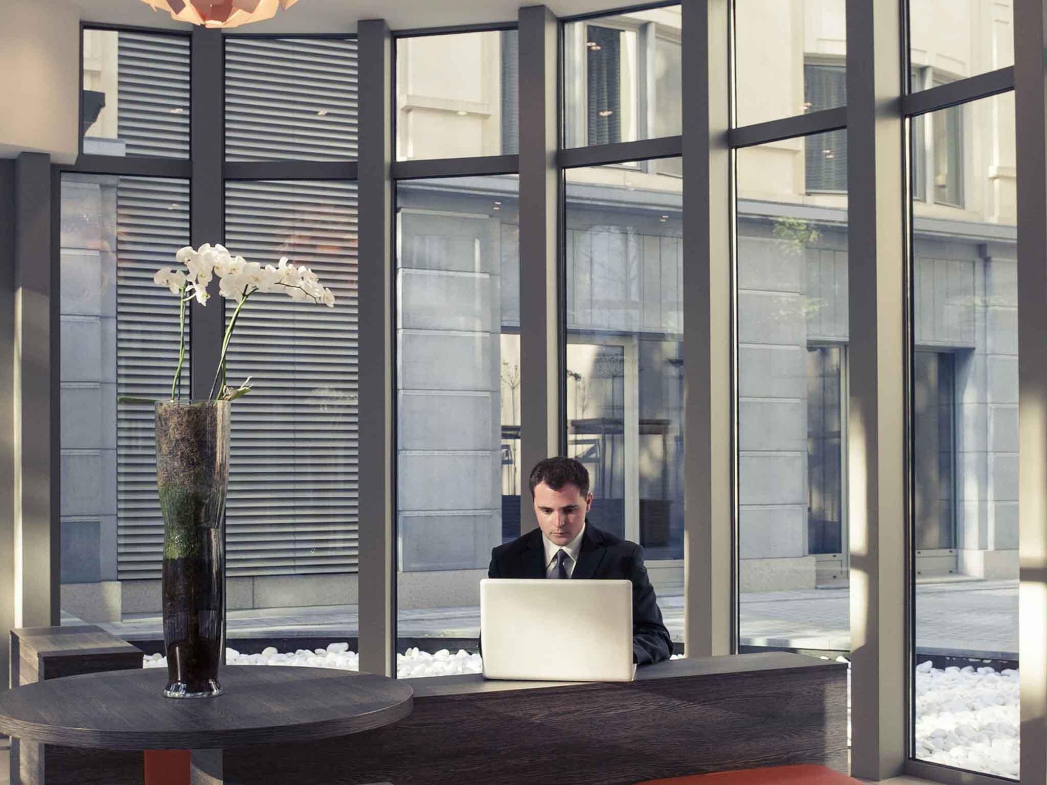 A man working at a desk with a laptop, large windows, and a vase with white flowers.