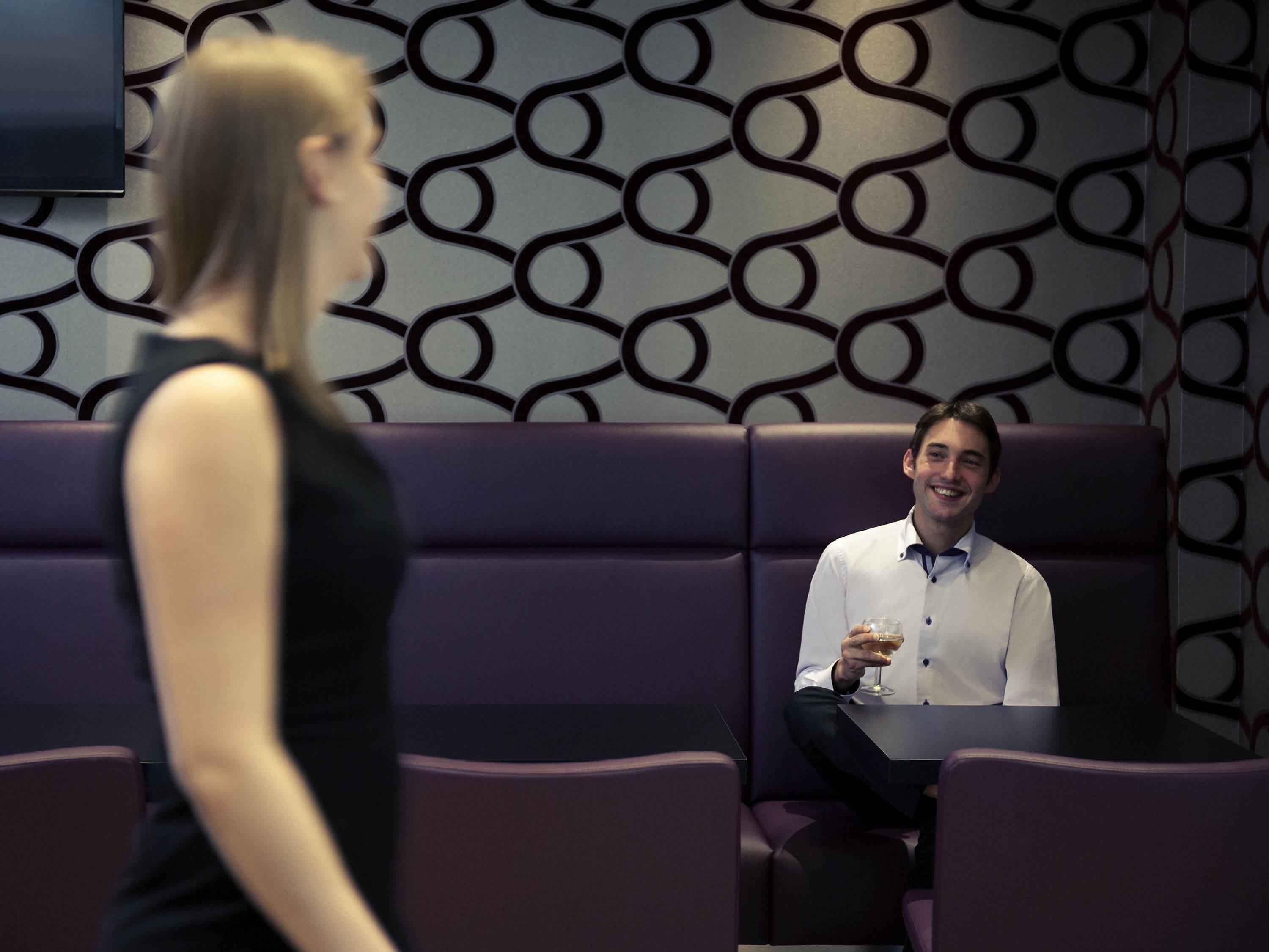 Ein modernes Interieur mit einer gemusterten Wand, einer Sitzecke mit violetten Polstern und einer Person, die ein Getränk hält.
