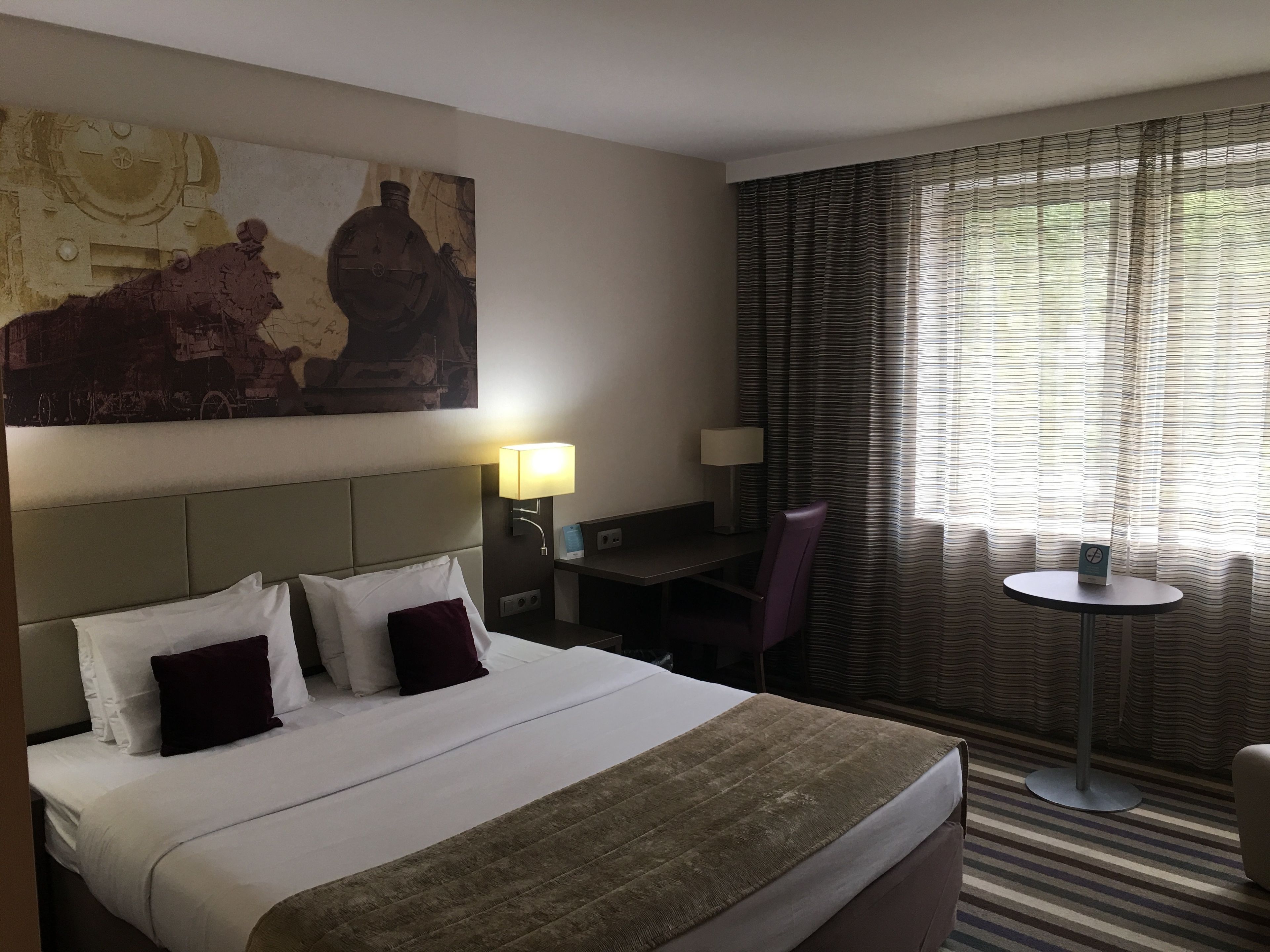 Habitación de hotel con una cama grande, un escritorio con silla, una mesita y una ventana con cortinas.